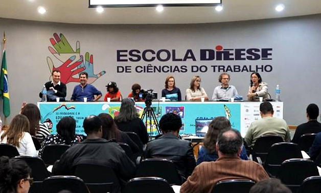 Foto muestra ocho panelistas. Detrás de ellos hay un banner que dice "Escola DIEESE de ciências do Trabalho"