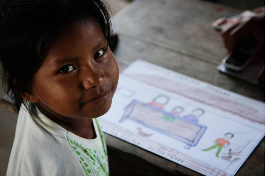 La foto muestra a una niña con rasgos indígenas, de aproximadamente 6 años de edad. Ella está sentada y viendo de lado hacia la cámara. Sonríe tímidamente. En el fondo desenfocado, está un dibujo coloreado sobre una mesa de madera oscura. Fin de la descripción.