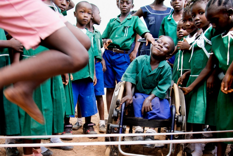 La foto muestra una aglomeración de niñas y niños negros alrededor de un niño en silla de ruedas, ellas y ellos están en la franja de edad entre los 5 y 7 años aproximadamente. Las niñas llevan un uniforme de falda y camisa verde con cuello blanco y los niños llevan un uniforme de camiseta tipo polo de color verde y una bermuda azul rey.